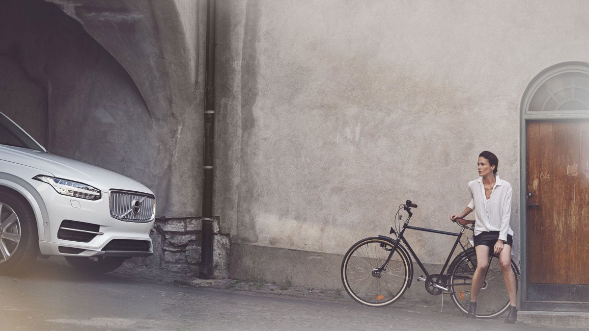 Nainen istumassa polkupyörän tarakalla katsomassa Volvo-autoa